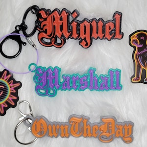 Puede incluir: Cinco llaveros con diferentes diseños. Un llavero presenta un sol negro y naranja con una cara sonriente. Otro llavero presenta una etiqueta de nombre morada y verde que dice "Marshall". Un tercer llavero presenta una etiqueta de nombre gris y naranja que dice "Own The Day". Un cuarto llavero presenta una etiqueta de nombre negra y naranja que dice "Miguel". El quinto llavero presenta una silueta de perro negra y neón.