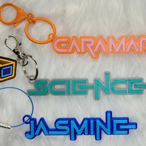 May include: Five colorful keychains with different designs.  The keychains include a pair of 3D glasses, a rainbow ice cream cone, and three names: "CARAMARIA", "SCIENCE", and "JASMINE".