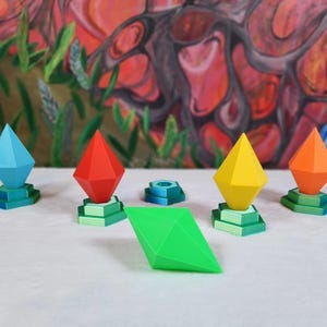 Può includere: Una collezione di oggetti geometrici a forma di diamante colorati. Gli oggetti sono in tonalità di blu, rosso, giallo e arancione, ciascuno su una base verde a più livelli. Una forma a diamante verde si trova di fronte agli altri.