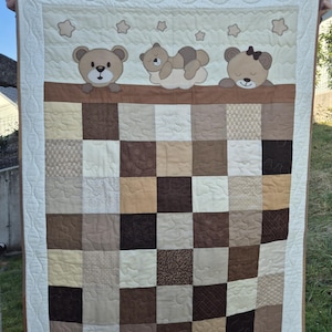 May include: A brown and beige patchwork quilt with a white border. The quilt features three embroidered teddy bears, two sleeping and one standing. The quilt is decorated with small stars.