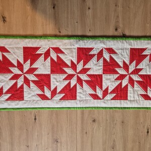 Pode incluir: Caminho de mesa acolchoado retangular com fundo branco e um padrão repetido de desenhos de estrelas vermelhas e brancas. O caminho de mesa tem uma borda verde. Aproximadamente 30 cm de largura e 76 cm de comprimento.