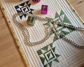 May include: A quilted Christmas table runner featuring geometric star patterns in red, navy, and green. Silver and red baubles, small gift boxes, and a silver beaded garland adorn the runner.