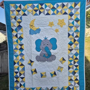 Könnte beinhalten: Eine weiße Babydecke mit einem blauen und gelben geometrischen Rand. Die Mitte der Decke zeigt einen grauen Elefanten mit blauen Ohren und einem blauen Rüssel, einen gelben Halbmond mit einem lächelnden Gesicht und drei gelbe Sterne mit lächelnden Gesichtern.