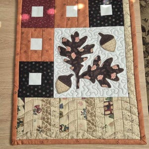 Può includere: Un runner da tavolo trapuntato con un design a patchwork marrone, arancione e bianco. Il centro presenta un'applicazione di foglie di quercia e ghiande in marrone e bianco.