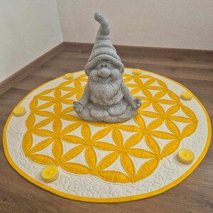 May include: A yellow and white flower of life mandala with a grey gnome statue sitting in the centre. The mandala is surrounded by small yellow candles.