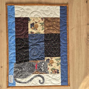 May include: A quilted table runner with a black cat design. The runner is made of various fabrics in shades of brown, blue, and white. The cat is in the lower right corner of the runner. The runner has a brown border with blue and white polka dots.
