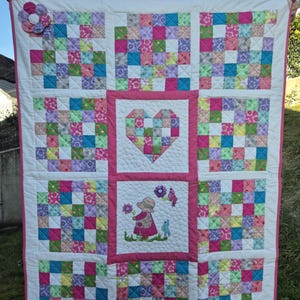 Peut inclure: Une courtepointe rose et blanche en patchwork avec un cœur, une fille avec une fleur et des oiseaux au centre. La courtepointe est faite de carrés de tissus différents dans des tons de rose, vert, bleu et violet.