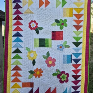 Può includere: Una trapunta bianca con un design floreale colorato. La trapunta è bordata con un bordo giallo e rosa. La trapunta presenta una varietà di fiori, foglie e forme geometriche.