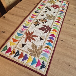 Può includere: Una tovaglia rettangolare trapuntata con un disegno centrale di foglie autunnali e ghiande nei toni del marrone e del beige. Il bordo è decorato con triangoli colorati. La tovaglia è lunga circa 152 cm.