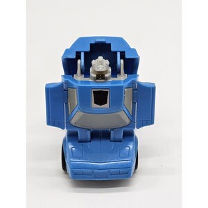 May include: A blue toy robot in a car configuration. The toy has a gray face and chest, with the robot's head visible. The car's doors are open, and the toy has black wheels. The toy has a black emblem on the chest.
