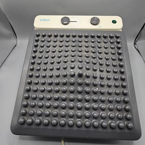 May include: A Vitec brand electronic foot massager featuring a dark gray rectangular base covered in a grid of raised circular nubs. The top control panel is off-white with a sliding switch, a circular dial, and a small green indicator light.