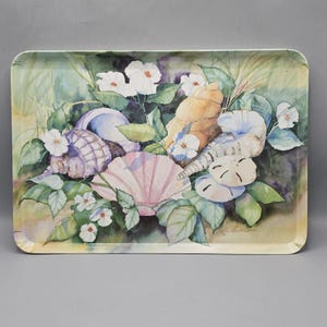 May include: A rectangular serving tray featuring a watercolor illustration of various seashells, including a pink scallop, a spiral conch, and two sand dollars, interspersed with white flowers and green leaves against a soft, muted background.