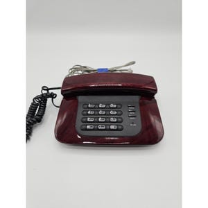 May include: A burgundy-colored, corded telephone with a black keypad and a wood-grain finish. The phone has a standard number pad, function buttons, and a coiled handset cord. The phone's design is a classic, landline style.