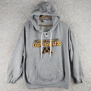 Puede incluir: Una sudadera con capucha y mangas largas en color gris jaspeado que presenta un cuello acordonado con cordones blancos. El frente muestra el texto UNIVERSITY OF MINNESOTA GOPHERS en amarillo y granate, junto a un gran logotipo M.