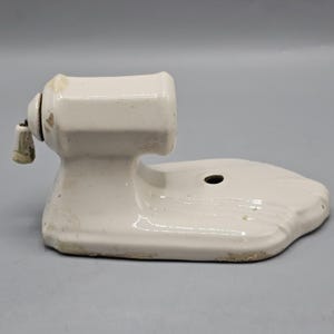 May include: A white ceramic wall-mounted soap dish featuring a horizontal cylindrical holder with a small attached knob and a flat, extended base with a single circular mounting hole. The surface has a glossy finish with minor signs of wear.