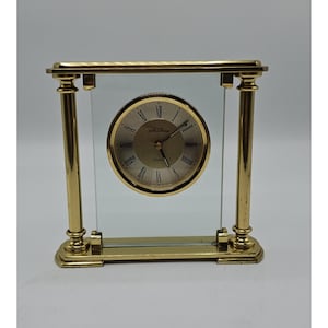 May include: A gold-toned quartz clock with a clear glass face and a gold frame. The clock face is circular with Roman numerals and gold hands. The clock is rectangular and sits upright.