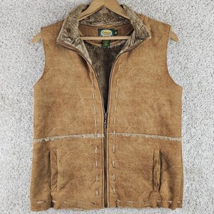 May include: A tan sleeveless vest with a textured faux suede exterior and a plush brown faux fur lining. The garment features a full-length front zipper, decorative whipstitch detailing along the seams and hem, and two vertical side pockets.