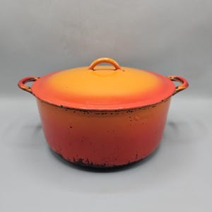 Peut inclure: Une cocotte en fonte avec un couvercle. Le pot est peint dans un dégradé d'orange et de rouge, avec une petite poignée sur le couvercle et deux poignées latérales. Le pot est conçu pour la cuisine.