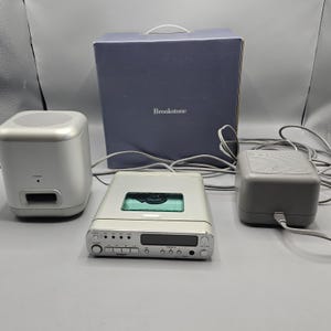 Könnte beinhalten: Ein silberner Brookstone CD-Player mit passendem Lautsprechersystem. Das System umfasst einen Subwoofer, zwei Lautsprecher und ein Netzteil. Der CD-Player hat ein Display und Bedientasten. Im Hintergrund befindet sich eine blaue Box mit dem Brookstone-Logo.