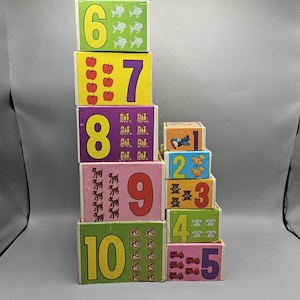 Pode incluir: Uma pilha de blocos de números vintage coloridos. Cada bloco apresenta um número de 1 a 10, juntamente com ilustrações de peixes, maçãs e outros objetos. Os blocos estão empilhados em uma torre, mostrando os números e as imagens.