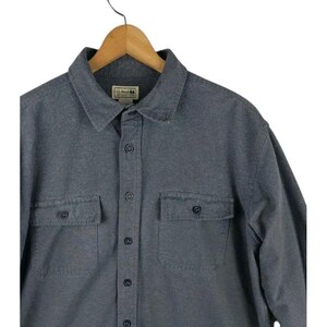 Puede incluir: Una camisa gris abotonada con dos bolsillos en el pecho y un cuello. La camisa está colgada en una percha de madera. El logotipo de la marca es visible en el cuello.