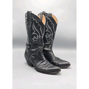 Op de afbeelding: Een paar zwarte cowboylaarzen met ingewikkelde zilveren stiksels. De laarzen hebben een textuur van leer en spitse tenen. De schachten zijn hoog en de hakken zijn schuin. De laarzen staan tegen een neutrale achtergrond.