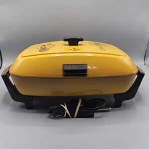 May include: A vintage yellow electric skillet with a black handle and base. The lid features decorative floral designs. The power cord is black and wrapped with a rubber band. The skillet is designed for cooking.