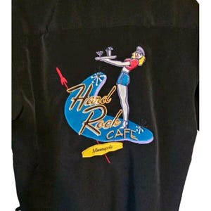 Pode incluir: Camisa preta com o logotipo Hard Rock Cafe nas costas. O logotipo apresenta uma garçonete carregando bebidas em um bumerangue azul, com o texto "Hard Rock Cafe Minneapolis".