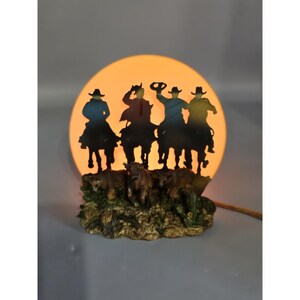 Pode incluir: Lâmpada decorativa com a silhueta de quatro cowboys a cavalo contra um fundo laranja brilhante. A base inclui figuras esculpidas de gado e plantas do deserto. A lâmpada tem um cabo de alimentação castanho.