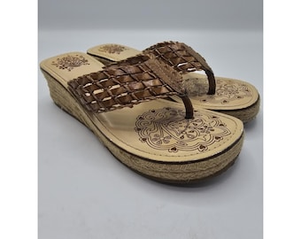 May include: A pair of wedge sandals with a woven brown strap and a beige footbed. The footbed features a decorative floral design. The wedge heel is wrapped in a natural-colored rope material.