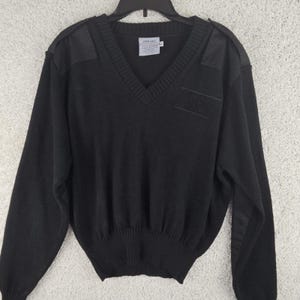 May include: Black V-neck jumper with shoulder patches and a ribbed hem. The jumper has a small label near the neckline and a patch on the chest. The long-sleeved jumper is hanging on a black hanger.