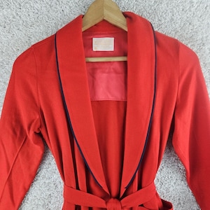 May include: A bright red robe featuring a shawl collar with dark navy blue piping trim. The garment includes a matching red fabric belt at the waist and a rectangular white label sewn into the inner neck area with visible black text.