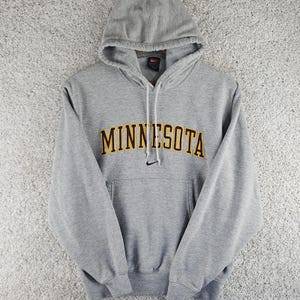 Puede incluir: Una sudadera con capucha de color gris jaspeado que presenta la palabra MINNESOTA en letras amarillas con un borde granate oscuro en el pecho. Un pequeño logotipo de Nike negro está bordado bajo el texto y cuelgan cordones blancos.