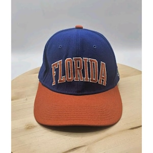 Peut inclure: Casquette de baseball bleue et orange avec le mot "FLORIDA" brodé sur le devant. La casquette a une visière incurvée et un bouton sur le dessus. Le chapeau est probablement fait d'un tissu durable.