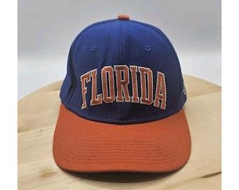 May include: A blue and orange baseball cap with the word "FLORIDA" embroidered across the front. The cap has a curved brim and a button on top. The hat is likely made of a durable fabric.