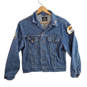 May include: A blue denim jacket with button fastenings, chest pockets, and a collared neckline. It has embroidered patches on the sleeves, including a flower and text. The jacket is hanging on a wooden hanger.