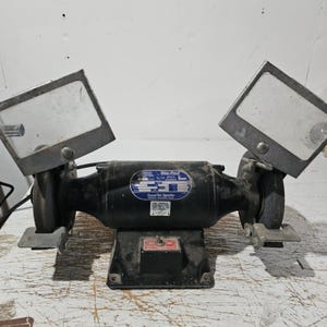 May include: A black Blue Point bench grinder with two grinding wheels and protective shields. The grinder has a rectangular base and a label with technical specifications. The shields are made of clear material and are mounted on adjustable arms.