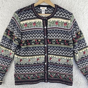 May include: A long-sleeve button-down cardigan featuring a dark base with repeating horizontal bands of zigzag patterns, floral motifs, and berry-like accents in shades of red, green, cream, and grey. The garment has a round neckline and dark buttons.