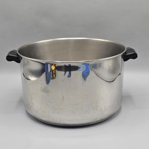 Può includere: Una pentola da cucina in acciaio inossidabile con un'ampia apertura e due manici neri. La pentola ha una forma cilindrica e una superficie lucida e riflettente. L'interno della pentola è vuoto. I manici sono attaccati ai lati.