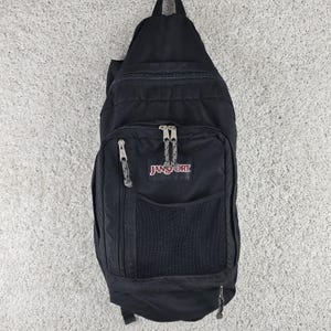 Puede incluir: Una mochila bandolera negra JanSport con un compartimento principal grande, un bolsillo frontal con cremallera y malla, y una cremallera lateral vertical. La bolsa incluye tiradores de cremallera de metal plateado con cordones grises.