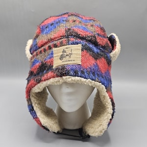 Peut inclure: Une chapka à motif géométrique multicolore dans les tons rouge, bleu et marron. Le bonnet est doté d'une doublure et de rabats d'oreilles en fausse fourrure crème. Un écusson rectangulaire beige affiche un motif et le texte MAD BOMBER.