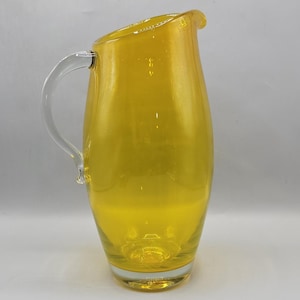 Pode incluir: Um jarro de vidro amarelo translúcido alto com um corpo liso e arredondado e uma pega de vidro transparente. O recipiente apresenta um topo largo e aberto com um bico vertedor proeminente, e a base é feita de vidro transparente.