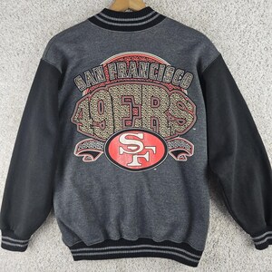 Könnte beinhalten: Ein anthrazitfarbenes Sweatshirt mit schwarzen Ärmeln sowie einem gerippten Kragen und Saum mit grauen Streifen. Auf der Rückseite befindet sich eine große Grafik mit dem Text SAN FRANCISCO 49ERS in strukturierter Optik über einem rot-weißen SF-Logo.