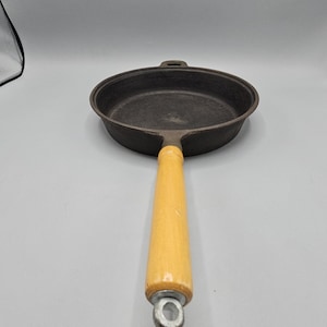 May include: A cast iron frying pan with a light wood handle. The pan is dark grey and has a round cooking surface. The handle is a light yellow colour and is attached to the pan with a metal ring.