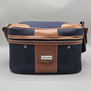 May include: A rectangular navy blue bag with brown leather-like trim and accents. The front features a central vertical brown panel with a silver-toned metal plaque embossed with the text JAGUAR. It includes a zippered closure and side straps.