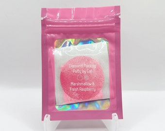 Könnte beinhalten: Ein rosa wiederverschließbarer Beutel mit einem runden rosa Behälter mit Diamant-Mal-Knete. Der Behälter hat ein weißes Etikett mit dem Text "Diamond Painting Putty by Cat" und "Marshmallow & Fresh Raspberry".