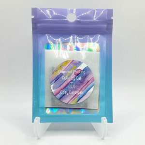 Puede incluir: Un paquete holográfico transparente que contiene una pegatina redonda con rayas diagonales de acuarela en rosa, azul y dorado. La pegatina dice "Diamond Painting Putty by Cat" y "Cosy Cardigan."