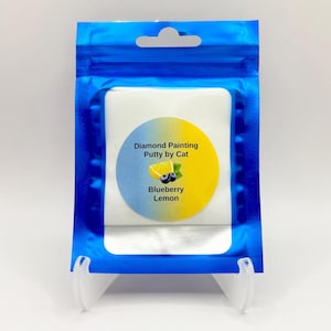 May include: A blue, resealable bag holding a white square package. The package has a circular sticker with "Diamond Painting Putty by Cat", a lemon and blueberry illustration, and the words "Blueberry Lemon".