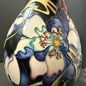 May include: A black ceramic vase with a floral design. The vase features large, stylized flowers in shades of white, blue, and yellow, with red and orange accents. The flowers are set against a black background, creating a striking contrast.