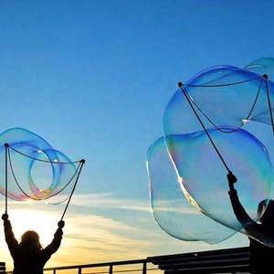 Peut inclure: Silhouette d'une personne créant de grandes bulles iridescentes avec une baguette contre un ciel bleu. Les bulles sont de tailles et de formes variées, reflétant la lumière du soleil. La scène se déroule au coucher du soleil.